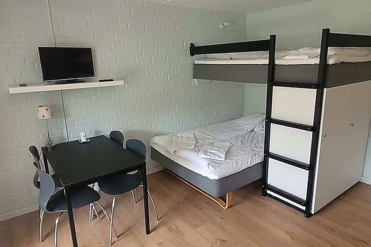 A room with a bunk bed, a desk with chairs, a television on a shelf, and a light-colored wall.