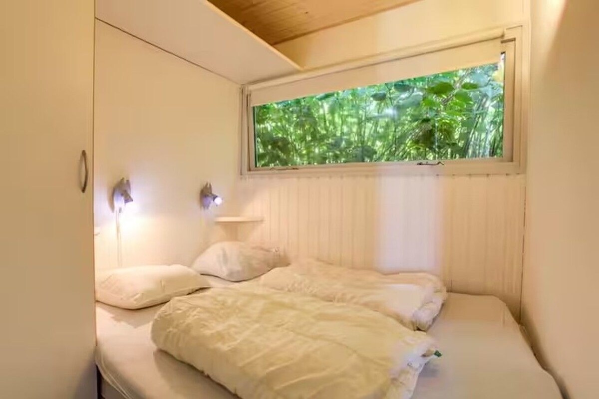 A bedroom with a bed, a window with a view of greenery, and a wall-mounted light fixture.