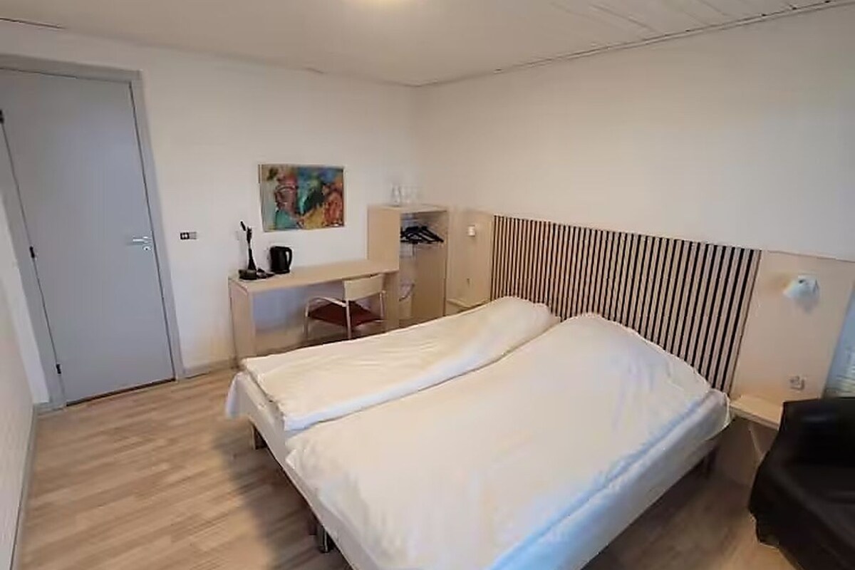 A hotel room with a double bed, a desk, a chair, a wardrobe, and a painting on the wall.