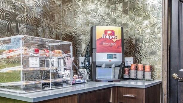 Un angolo caffè con una macchina da caffè Folgers Select Brew, un contenitore di plastica trasparente con diversi oggetti e una parete decorata sullo sfondo.