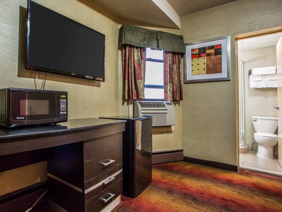 Habitación de hotel con televisión de pantalla plana, microondas, refrigerador pequeño, equipo de aire acondicionado, escritorio de madera con cajones, alfombra estampada y un cuadro en la pared.