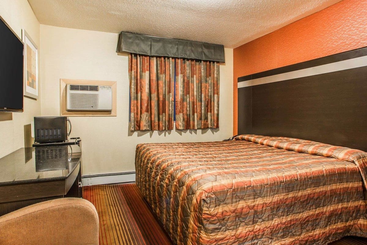 A hotel room with a bed, a desk with a chair, a television, and an air conditioning unit.