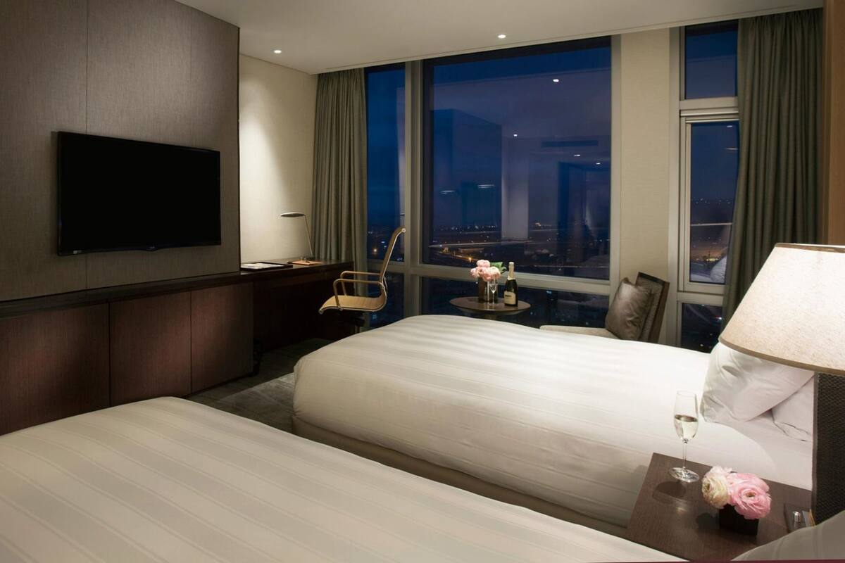 Habitación de hotel con dos camas, un televisor de pantalla plana montado en la pared, un escritorio con una silla, una ventana con vistas a la ciudad y una mesita de noche con una lámpara y flores.