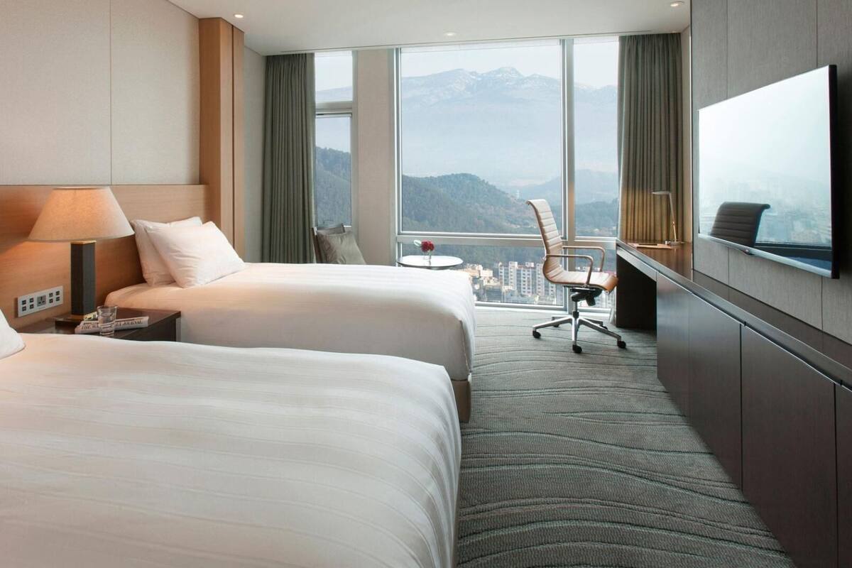 Habitación de hotel con dos camas, un escritorio, una silla, un televisor y un ventanal con vista a la montaña.