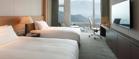 A hotel room with two beds, a desk, a chair, a TV, and a large window with a mountain view.