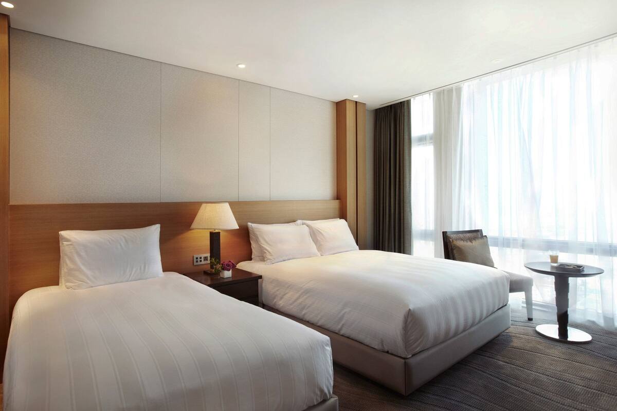 Habitación de hotel con dos camas, una mesita de noche con lámpara, una pequeña mesa y vistas a la ciudad a través de amplios ventanales.