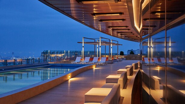 A rooftop pool area with lounge chairs and a glass balustrade overlooking a cityscape at dusk.