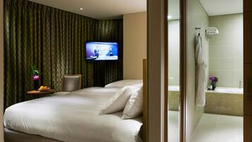 Habitación de hotel con dos camas, un televisor, una mesita con un florero y fruta, y un baño visible a través de una puerta abierta.