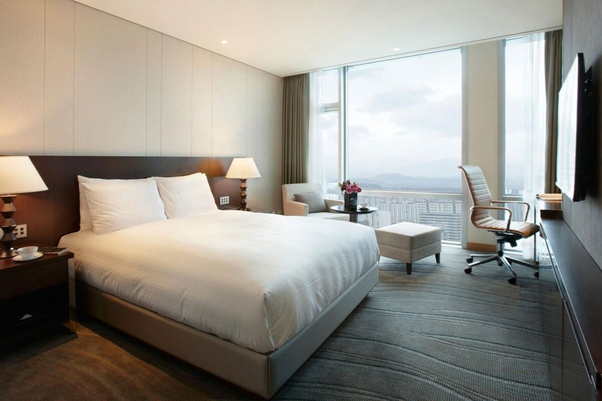 A modern hotel room with a large bed, two bedside lamps, a small table with a cup and a remote control, a chair, and a view of the city through large windows.