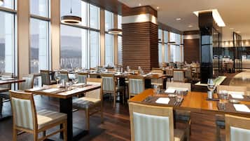 A modern restaurant with multiple dining tables and chairs, large windows offering a city view, and a high ceiling with recessed lighting.