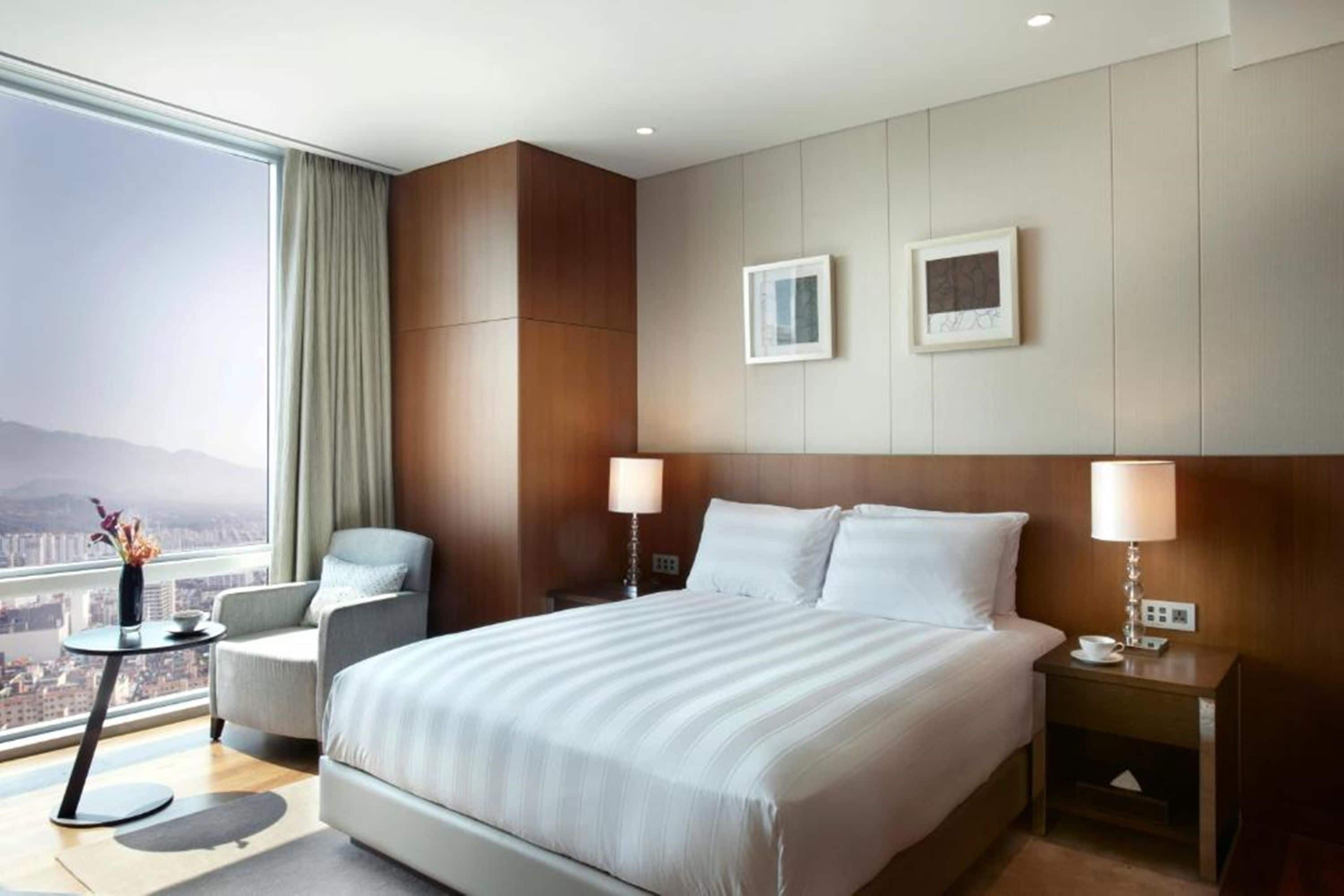 A modern hotel room with a large bed, two bedside tables, a chair, a view of the city, and two framed pictures on the wall.