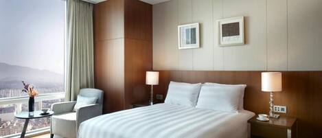 A modern hotel room with a large bed, two bedside tables, a chair, a view of the city, and two framed pictures on the wall.