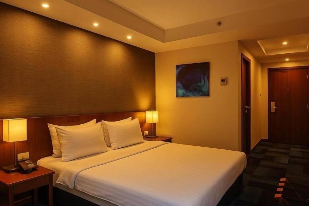 A hotel room with a large bed, two bedside tables with lamps, a painting on the wall, and a door leading to another room.