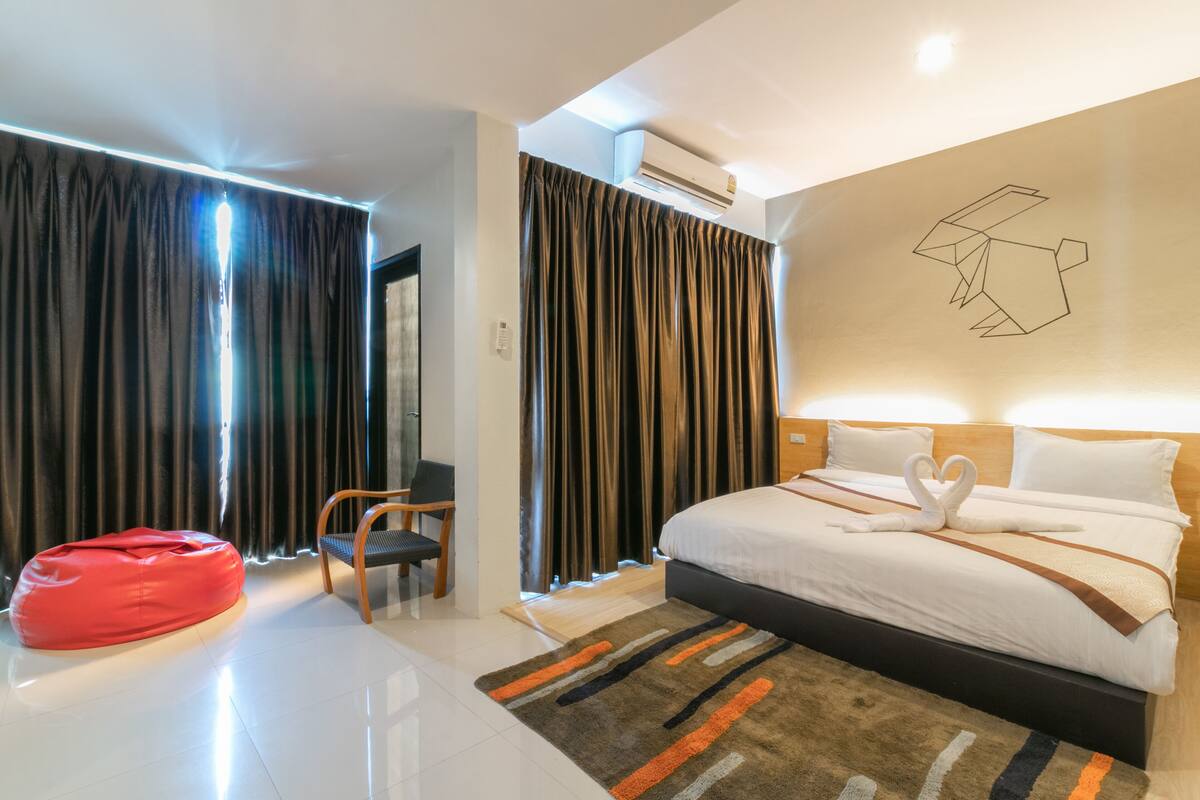 A modern hotel room with a large bed, a red bean bag chair, a wooden chair, and a patterned rug.