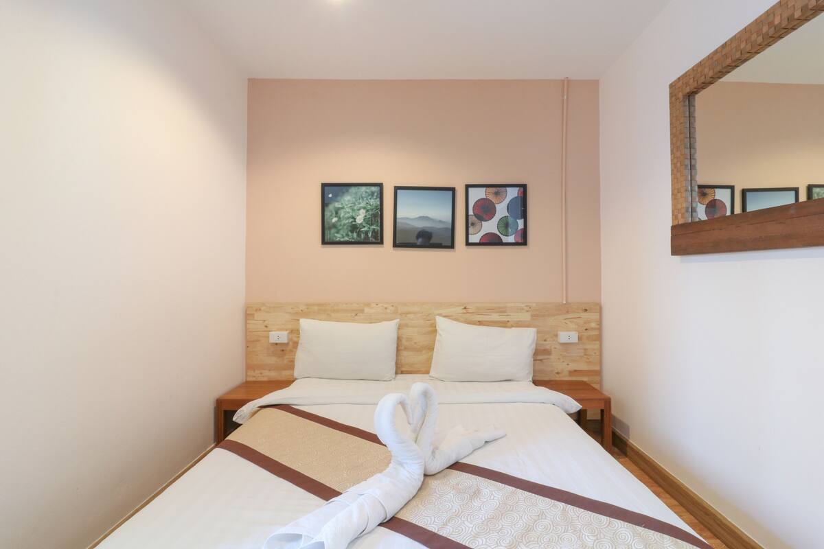 A hotel room with a wooden headboard, a bed with white linens, and a towel artfully placed on the bed. There are three framed pictures on the wall above the bed.