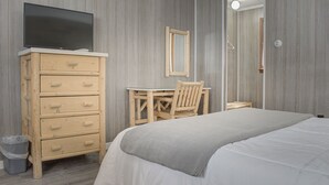 A hotel room with a bed, a wooden dresser with drawers, a television on top, a desk with a chair, and a mirror on the wall.