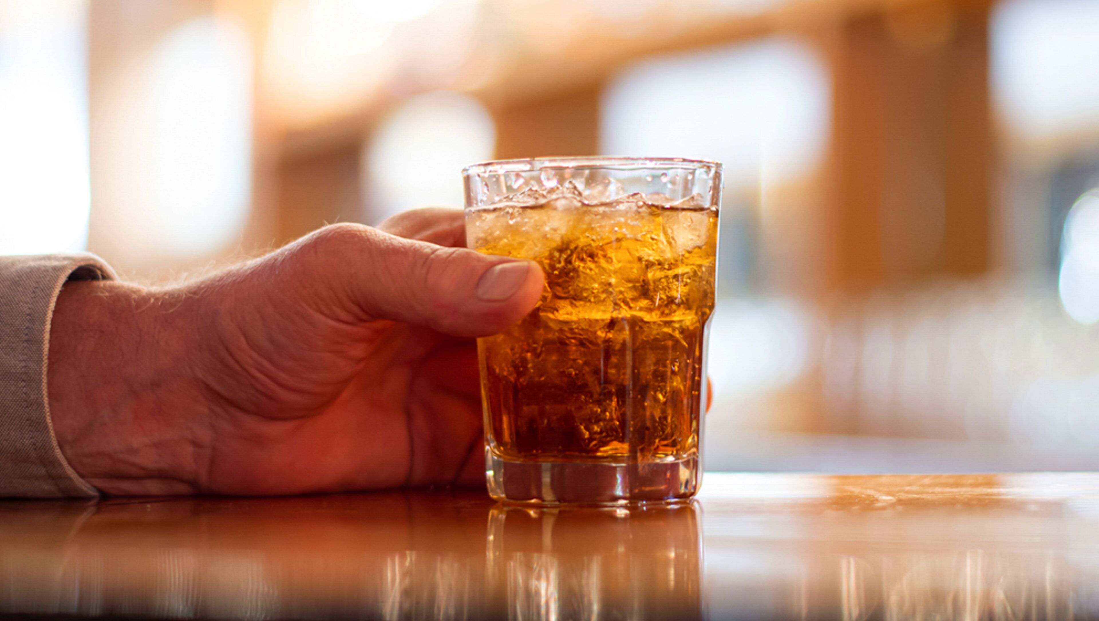 En hånd holder et glas med en ravfarvet drik, muligvis te eller whisky, med isterninger.