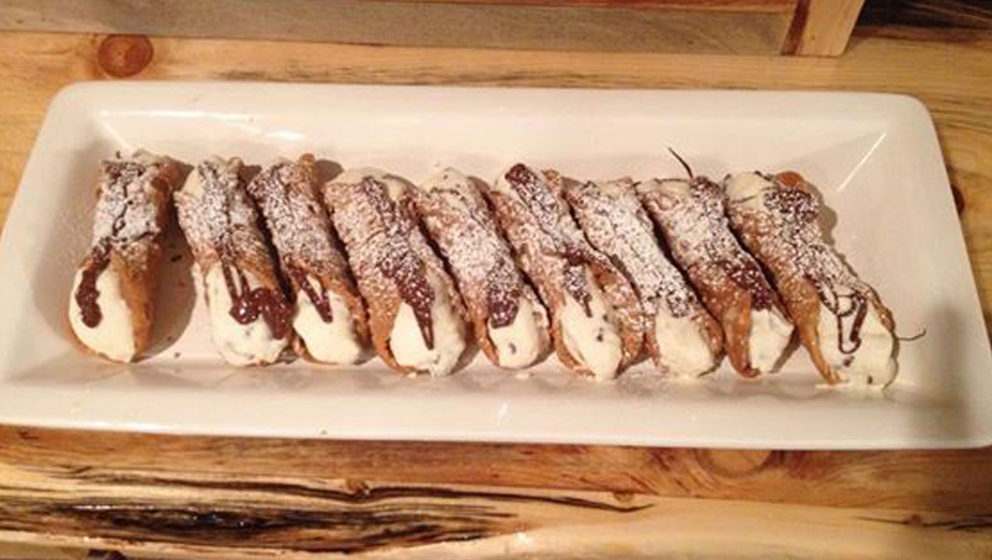 A white rectangular plate with a row of cream-filled pastries dusted with powdered sugar and drizzled with chocolate.