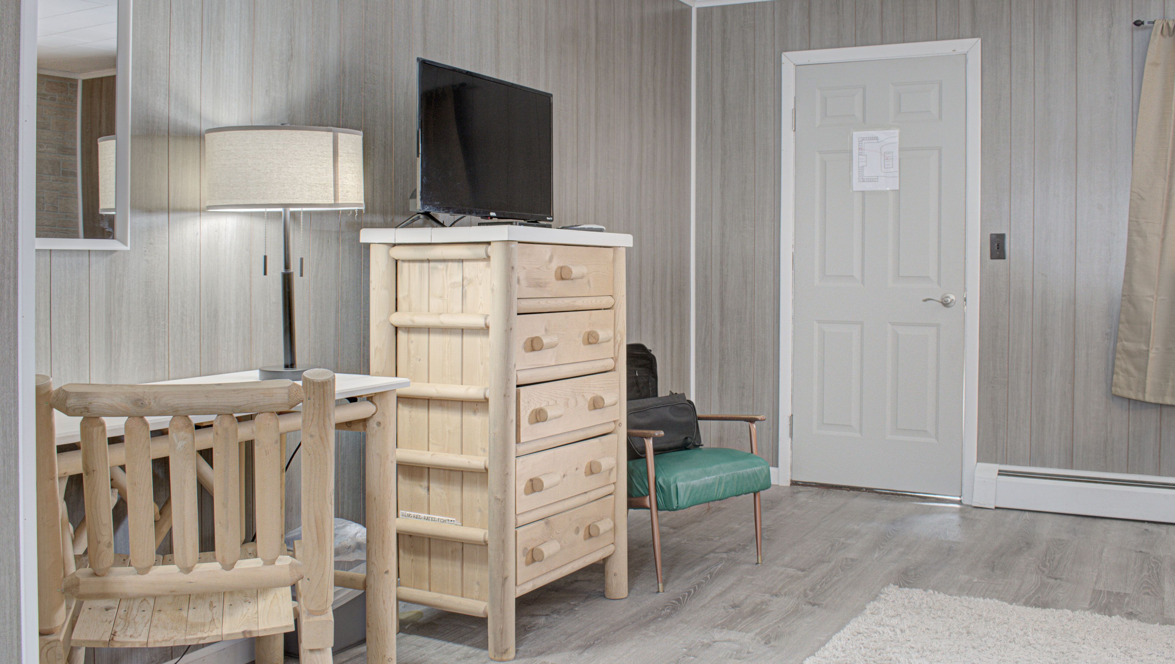 A room with a wooden dresser, a television on top, a desk with a chair, a lamp, and a door with a sign.