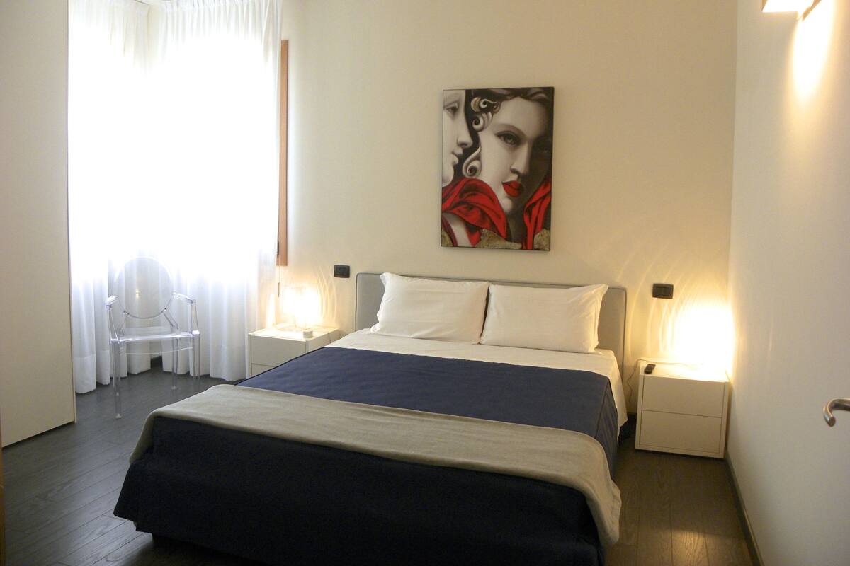 A hotel room with a bed, two bedside tables, a nightstand, a chair, and a painting on the wall.