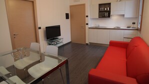 A modern living room with a glass dining table, a red sofa, and a kitchen area with white cabinets and a microwave. - Venice Apartments Dante (Mestre)