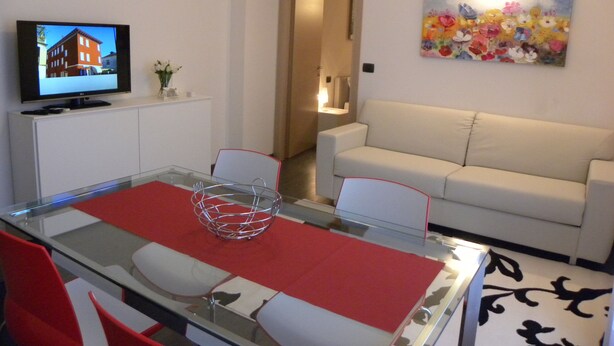A modern living room with a glass dining table, red placemat, and a beige sofa. A flat-screen TV is mounted on the wall, and a colorful floral painting is on the wall. - Venice Apartments Dante (Mestre)