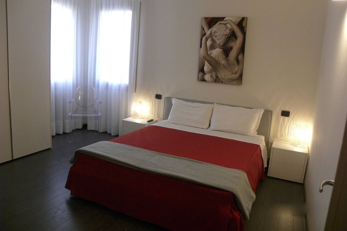 A hotel room with a large bed, two bedside tables with lamps, a painting on the wall, and a window with curtains.