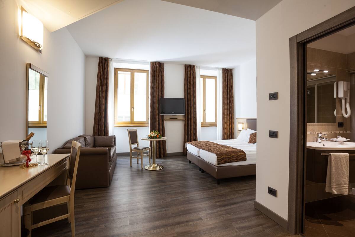 A hotel room with a bed, a desk with a chair, a TV, a small table with a flower arrangement, and a bathroom with a sink and mirror.