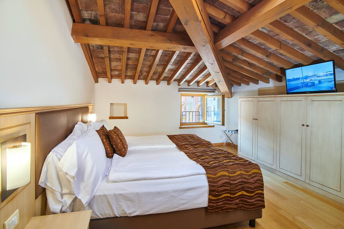 A bedroom with a large bed, wooden beams on the ceiling, a television on a cabinet, and a window with a view.