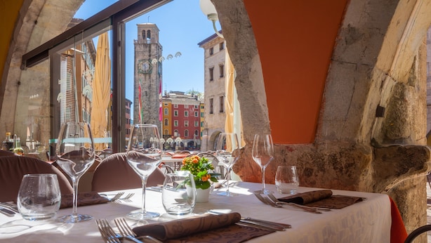 Mesa de comedor dispuesta con copas de vino, cubiertos y un jarrón con flores, con vistas a un casco antiguo con una torre de reloj.