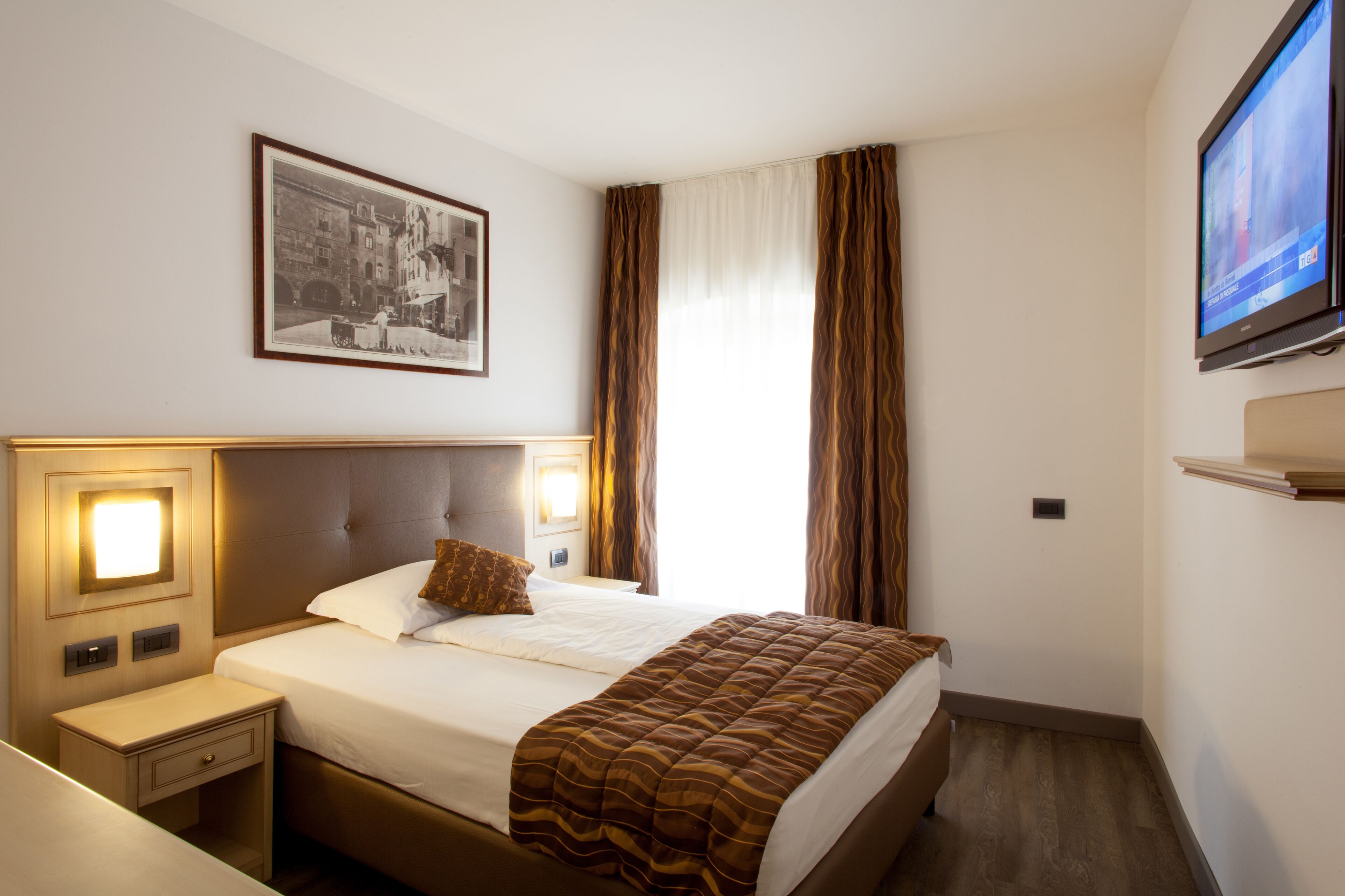 Una habitación de hotel con una cama, una mesita de noche, un cuadro enmarcado en la pared, un televisor y cortinas.