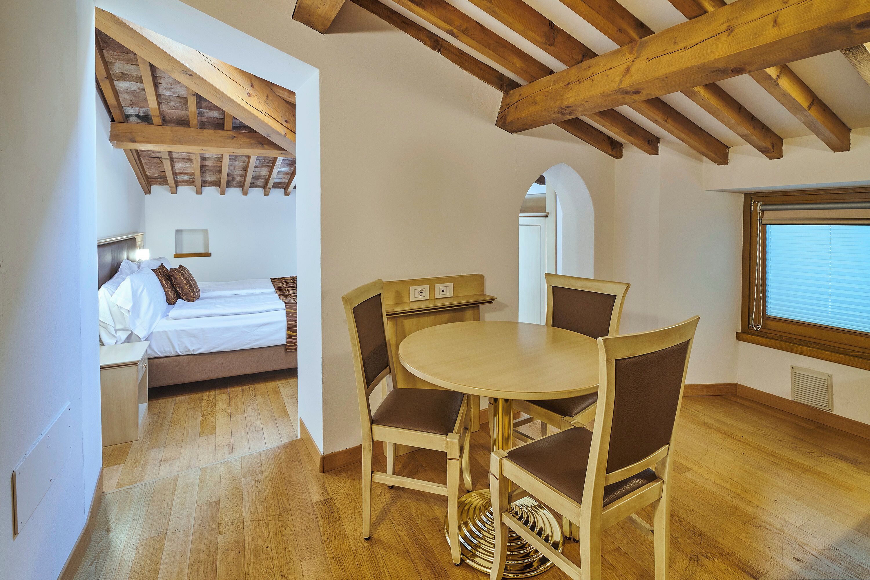 Una habitación con cama, una mesa redonda con sillas, vigas de madera en el techo y una ventana con persianas.