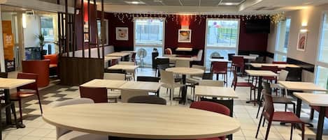 A spacious indoor dining area with multiple round tables and chairs, large windows, and a modern ceiling design.