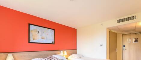 Une chambre d’hôtel avec deux lits, un mur d’accent rouge, un tableau encadré et une porte donnant sur une autre pièce.