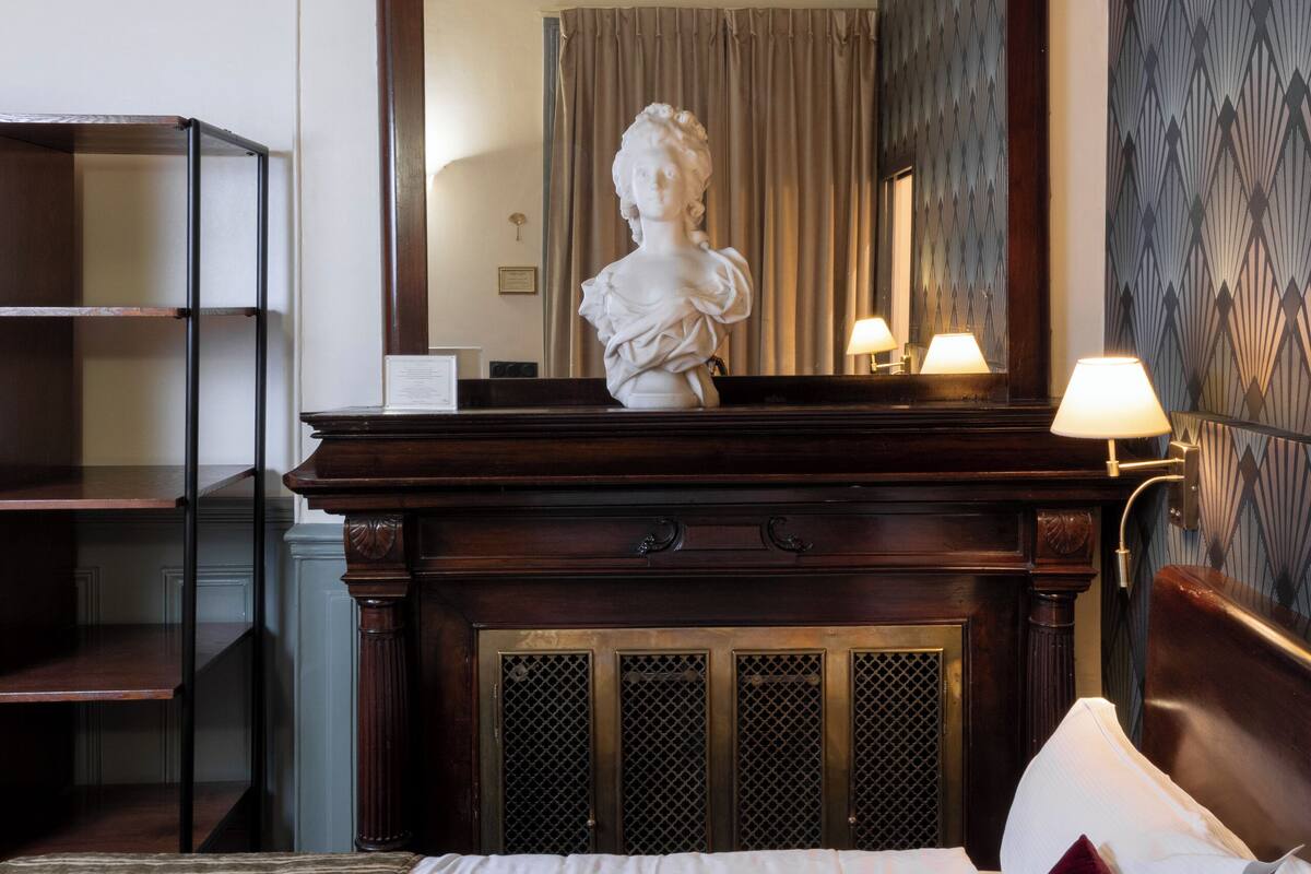 A hotel room with a bed, a wooden dresser, a mirror, and a bust sculpture on the dresser.