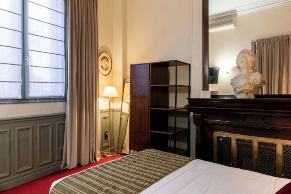 A hotel room with a bed, a nightstand, a lamp, a mirror, and a statue.