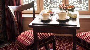 A dining table with a teapot, cups, and a basket of bread, set against a window with a view of a balcony and red flowers. - Hotel Langlois (Paris)