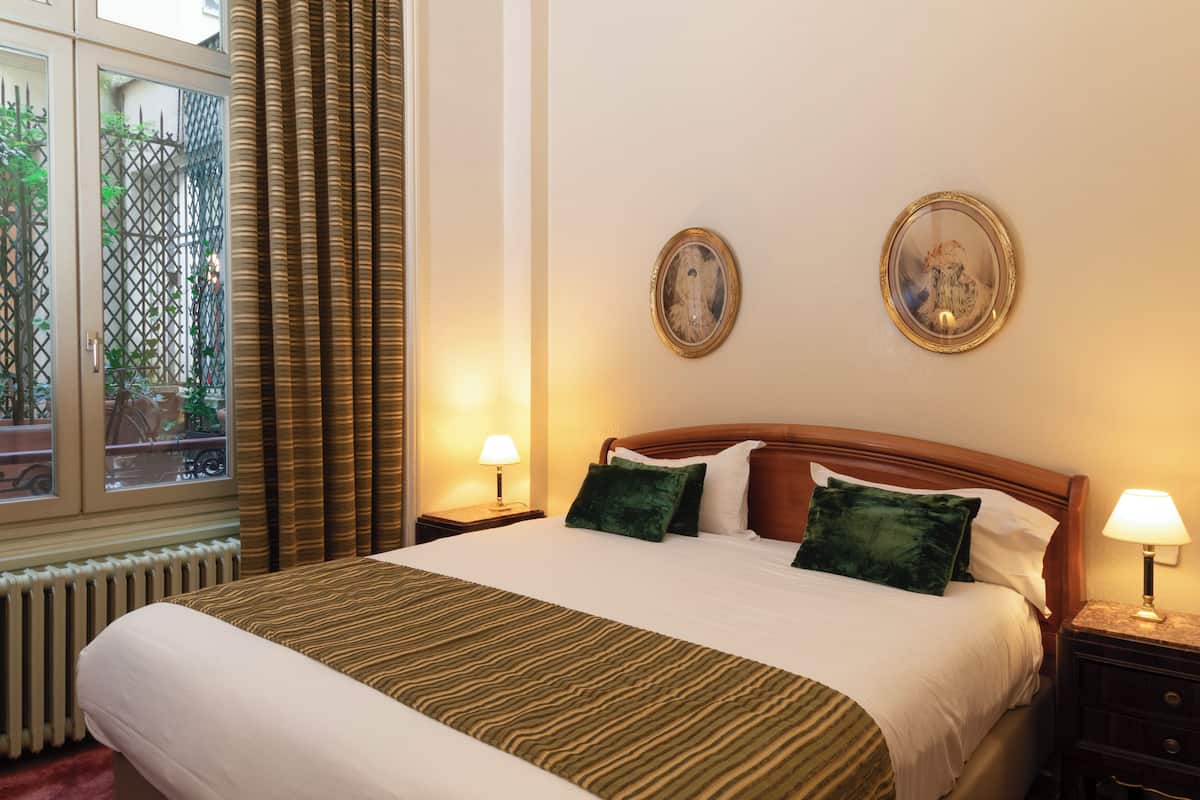A hotel room with a large bed, two bedside tables with lamps, a window with curtains, and two framed pictures on the wall.