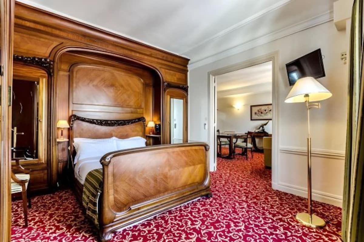 A hotel room with a large wooden bed, a red patterned carpet, a television, and a lamp.