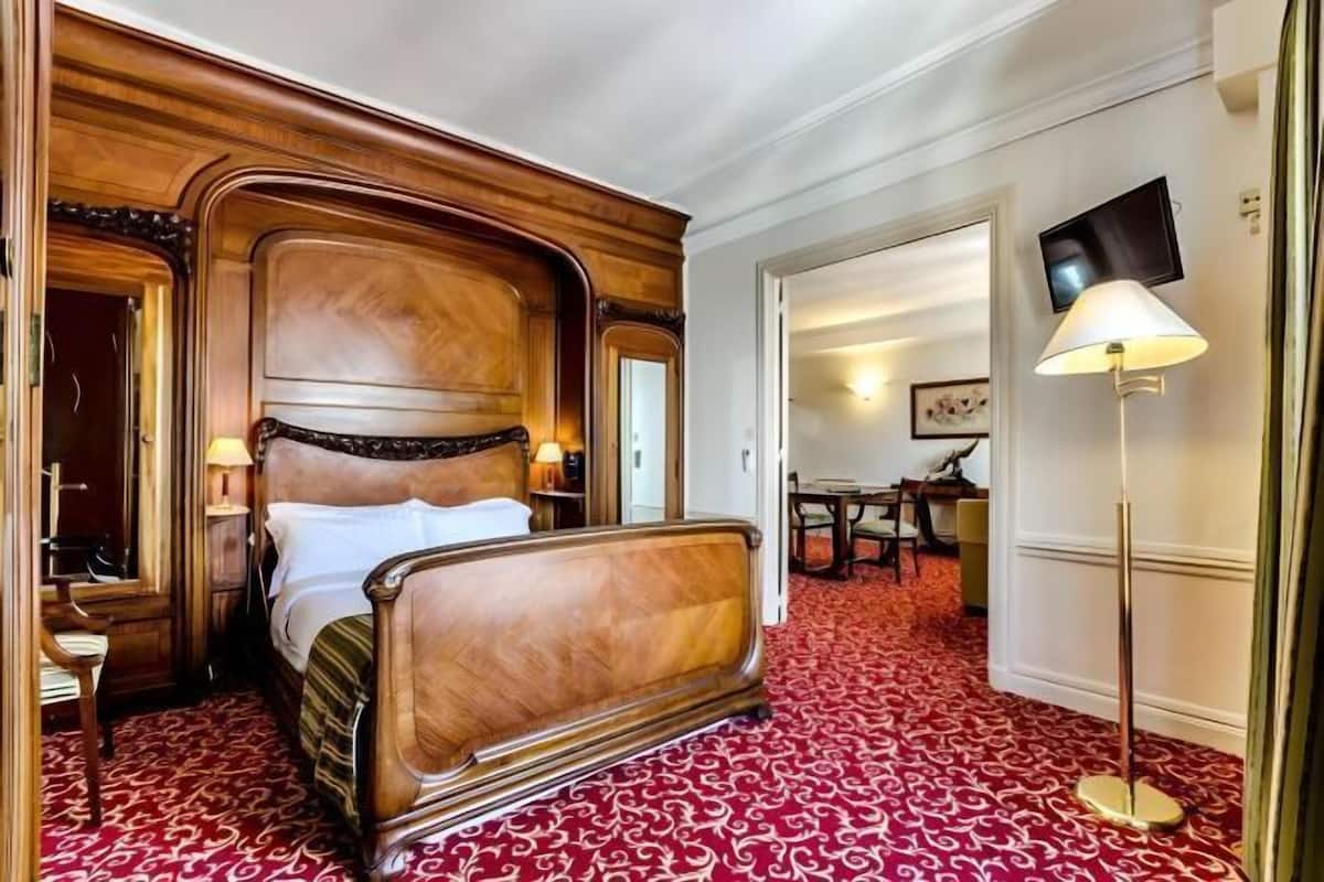 A hotel room with a large wooden bed, a red patterned carpet, a television, and a lamp.