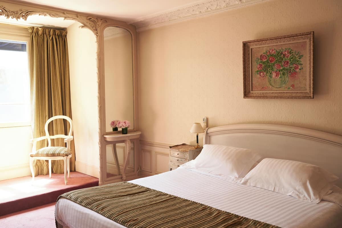 A hotel room with a large bed, a chair, a small table with flowers, and a painting on the wall.