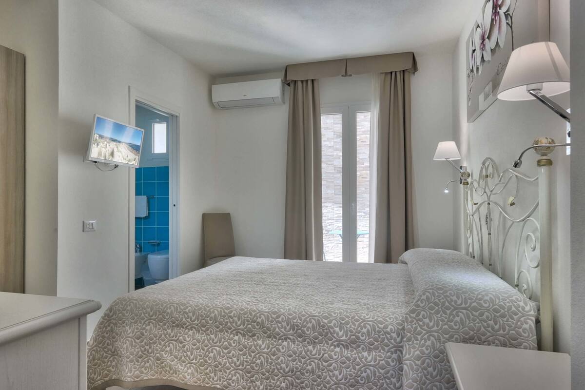 A bedroom with a bed, a TV, a bathroom with blue tiles, and a window with curtains.