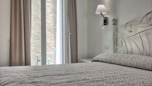 A bedroom with a bed, a nightstand, a wall-mounted lamp, and a view of the outside through a window with curtains. - Smart Suite & Apartments (Santa Teresa di Gallura)