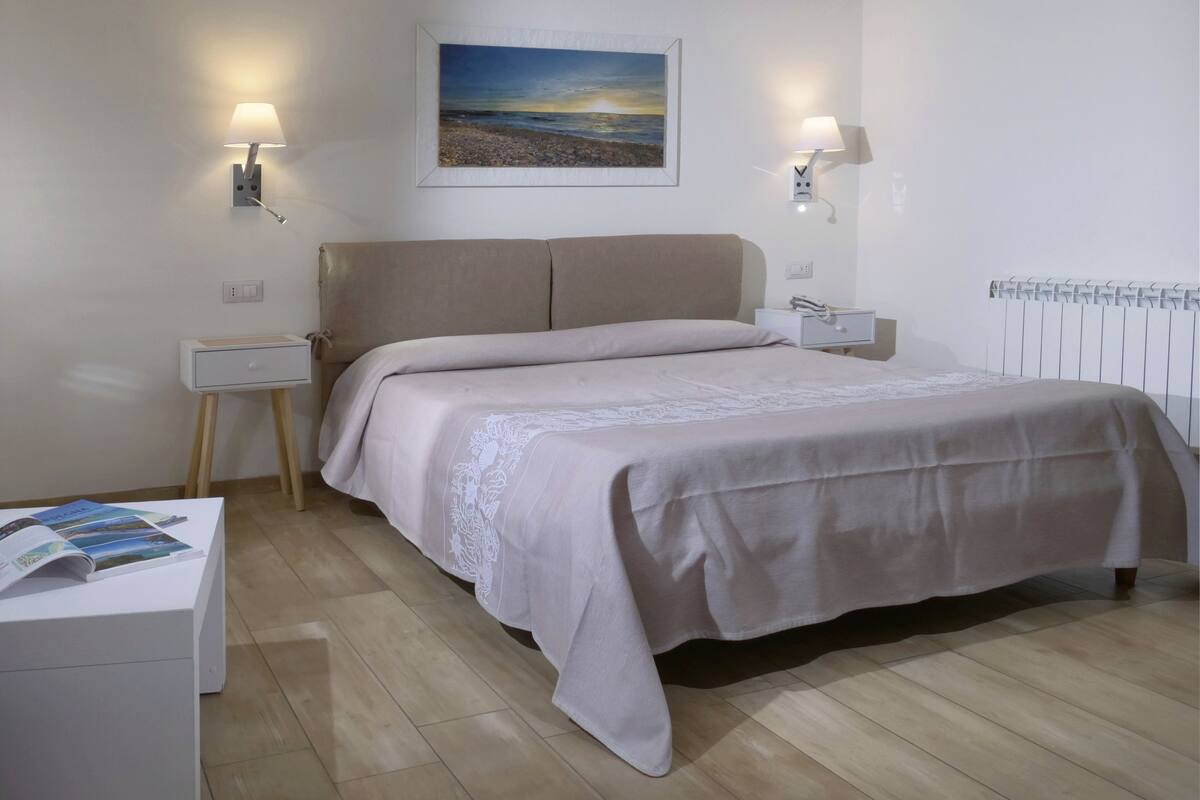 A bedroom with a bed, bedside tables, a nightstand, a wall-mounted lamp, a framed picture, and a radiator.