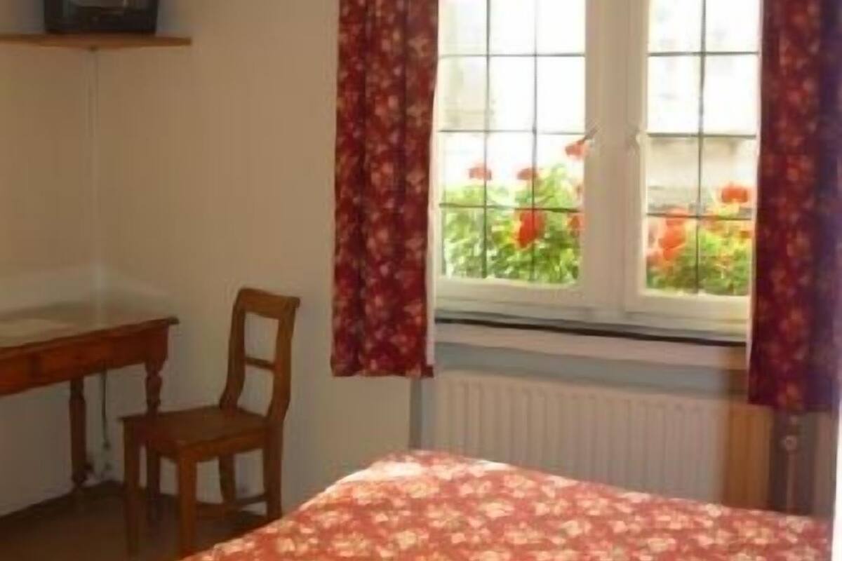 Ein Schlafzimmer mit Bett, Stuhl, kleinem Tisch, Fernseher auf einem Regal und einem Fenster mit Vorhängen.