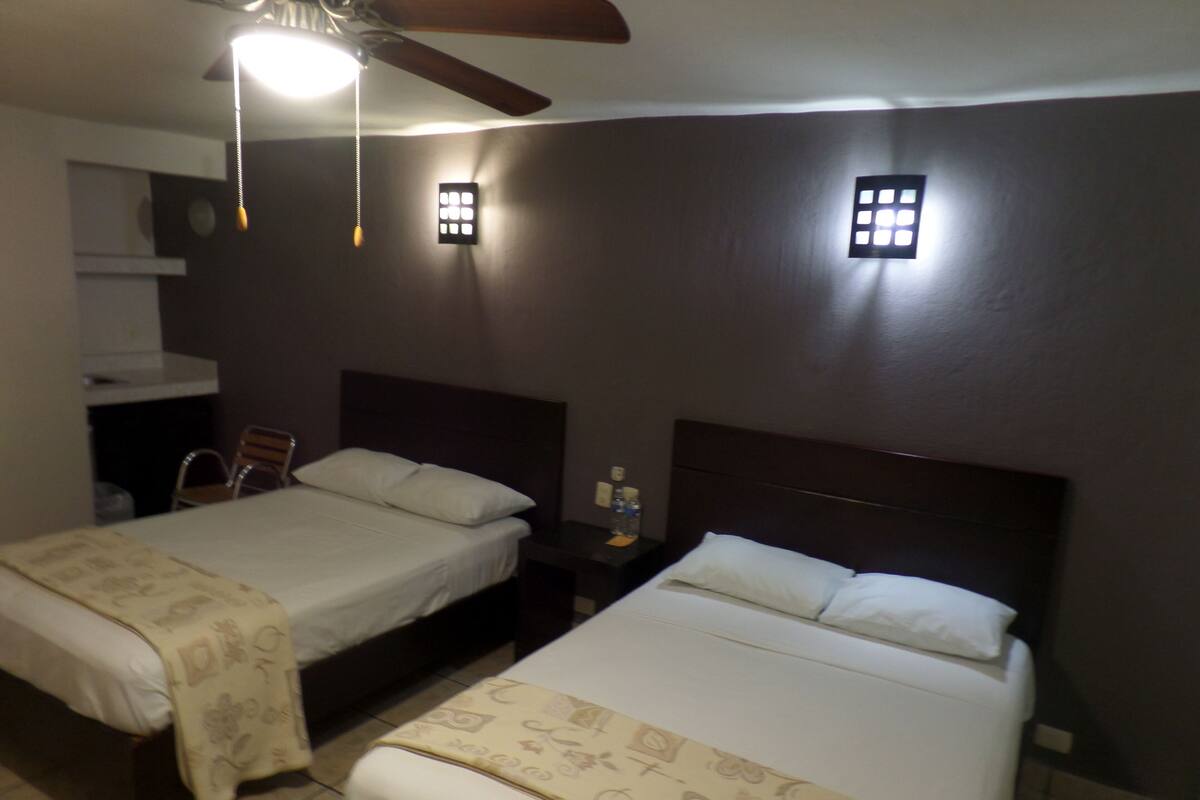 Quarto de hotel com duas camas, um ventilador de teto, uma mesa de cabeceira com abajur e uma pequena janela com vista para o exterior.