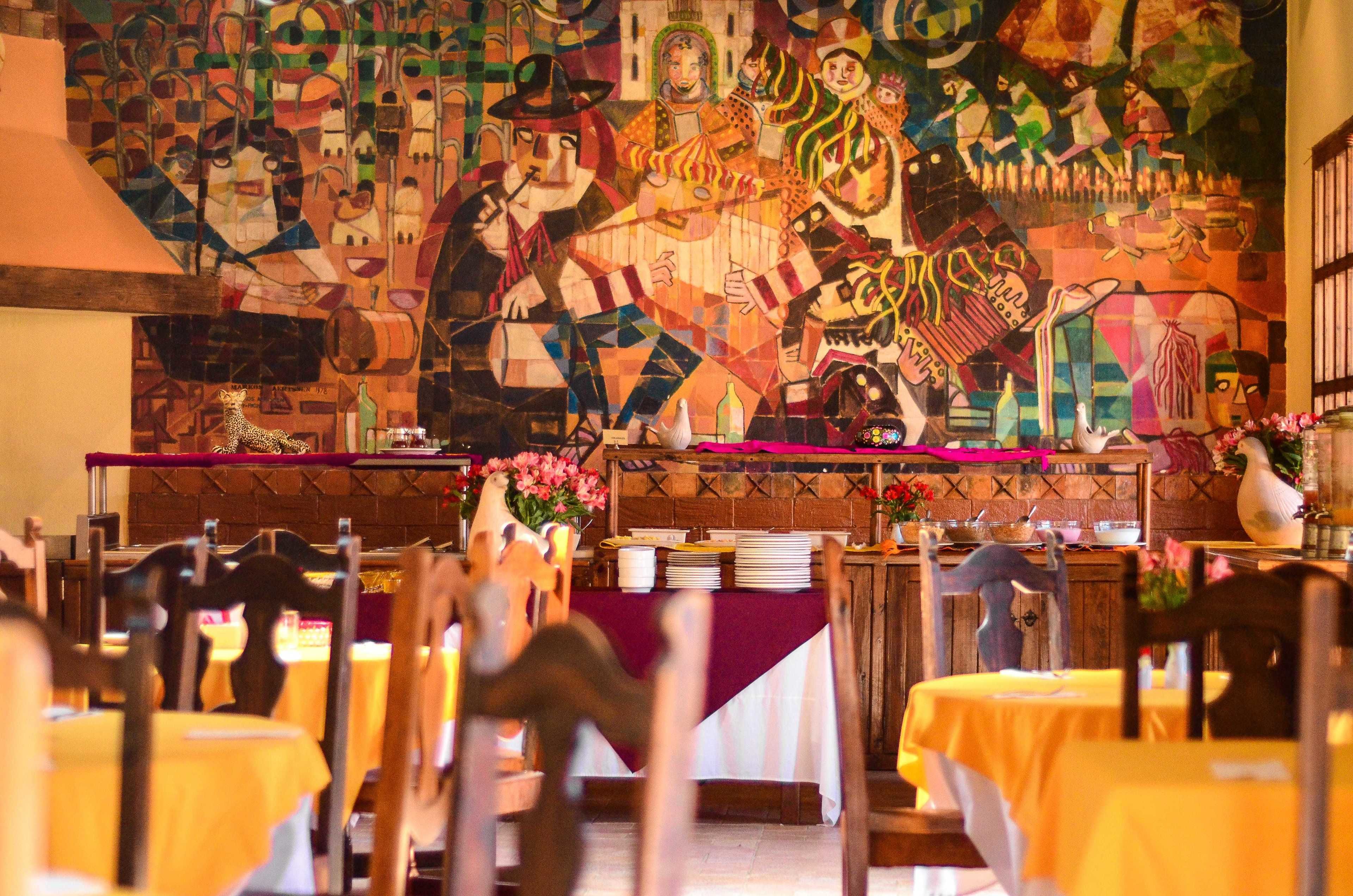 Une salle à manger avec des tables dressées, une fresque murale et un comptoir en bois recouvert d’un tissu rouge.