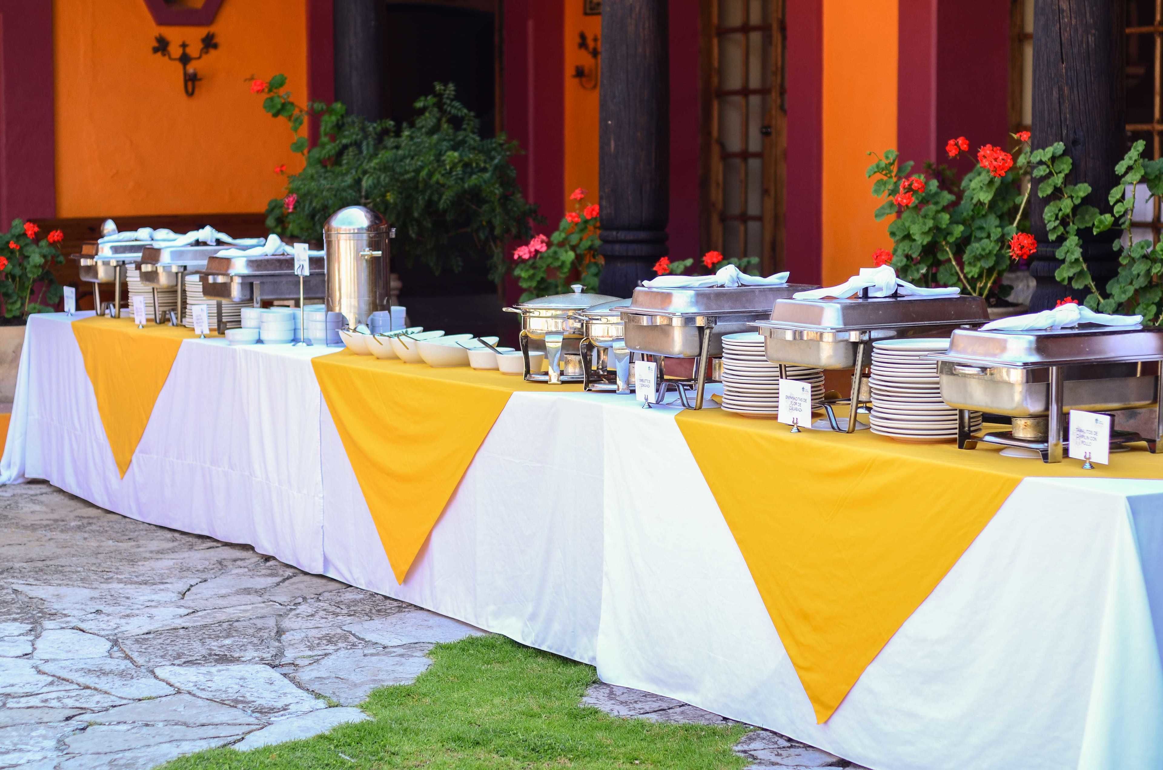 Une table de buffet installée à l’extérieur, avec plusieurs points de service, entourée de plantes en pot et bordée par un bâtiment aux murs orange et rouges.