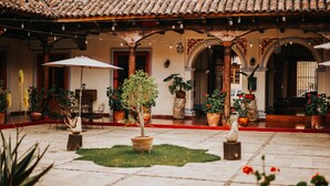 Un patio con suelo de baldosas, plantas en macetas, mobiliario exterior y una estructura de techo a la vista.