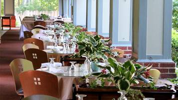 Une longue table à manger, entourée de chaises, agrémentée de verres et de plantes en pot, placée sous une zone abritée avec des fenêtres.